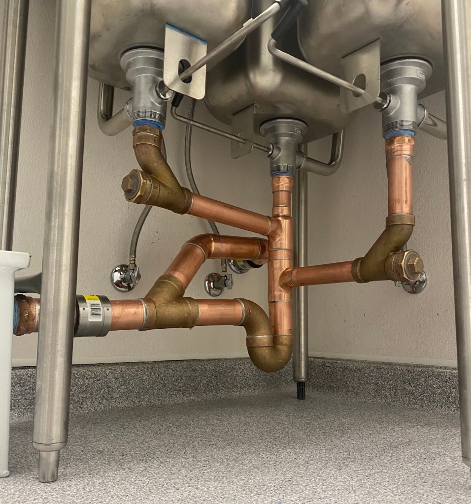 A close-up view of plumbing under a sink, featuring copper and metal pipes connected to a stainless steel sink.