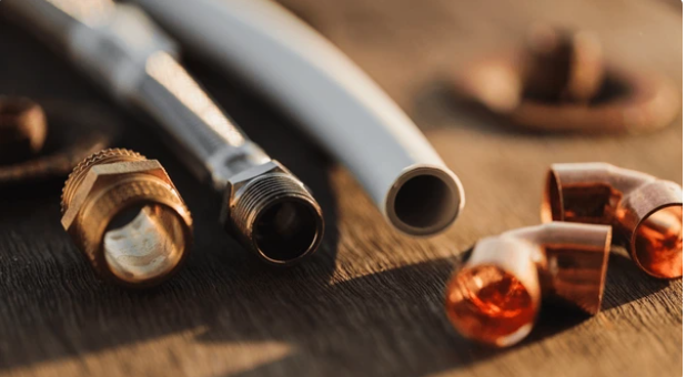 Close-up of plumbing materials including a metal hose, a white plastic pipe, and several copper fittings on a wooden surface.