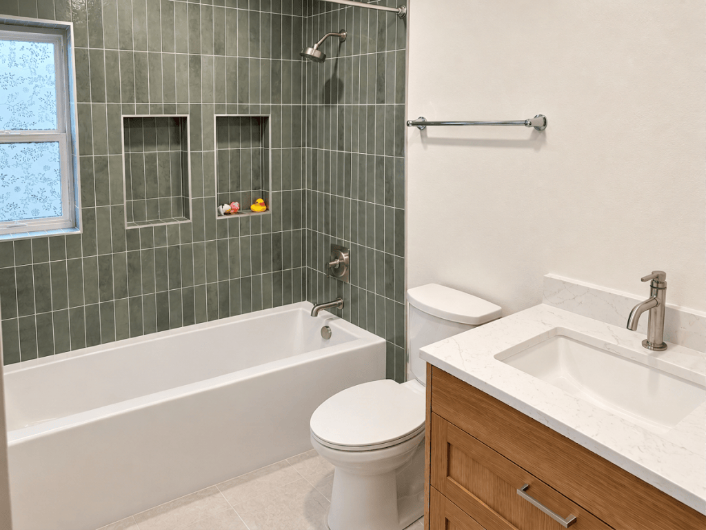 A modern bathroom featuring a white bathtub, green tiled walls, and a stylish vanity with a sink. Two wall niches display small colorful bath toys, and a window is visible to the left.