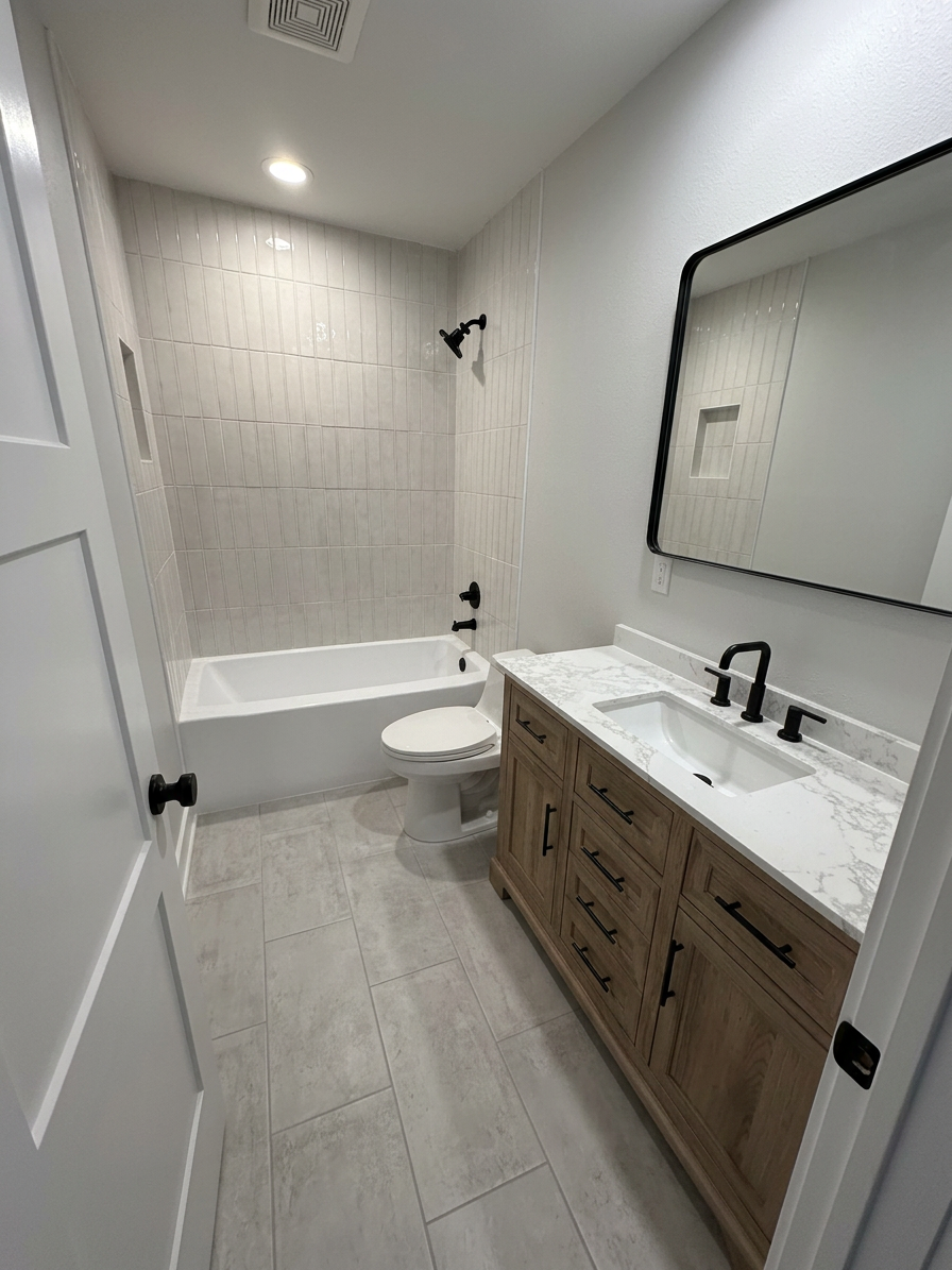 IMG_1820 Modern bathroom featuring a white bathtub, toilet, and a wooden vanity with a marble countertop and black fixtures. The walls are tiled with light-colored tiles, and there is a large mirror above the sink.